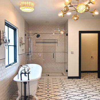 Bathroom design with horizonal grey subway tiles half way up the walls and in shower walls. Custom black and white mosaic flooring, and antique look for white tub with black hardware. Shower at the back of the room has black and white floor and niche details. Installation by GWH Flooring in Wimberley.
