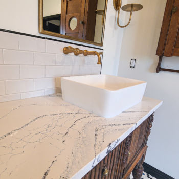 Custom designed bathroom with black and white marble countertop on antique vanity with white basin. Brass hardware, lighting, and mirror frame. Custom mosaic black, white, and blue penny tile flooring. Installation by GWH Flooring in Driftwood, Texas