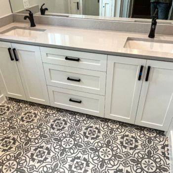Bathroom with grey quartz countertops for double sink vanity that has white cabinets. Black hardware. Floor is black and white decorative porcelain tile. Installation by GWH Flooring.
