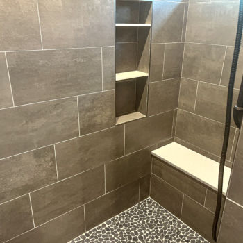 Shower with large grown ceramic tiles and white grout for walls. Stripe of brown and white mosiac squares from floor to ceiling. Brown pebble floor. White siltestone seat and niche shelves. Installation by GWH Flooring and countertops in Wimberley.