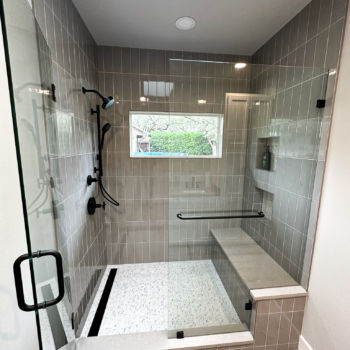 Grey subway tiles installed vertically for shower walls. Grey Silestone shower bench, mozaic shower floor with black hardware. Glass doors. Grey wood grain floor tiles. Installation by GWH Flooring in Wimberley.