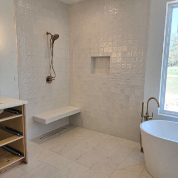 White square zia tiles for shower walls with floating bench above hidden drain. White rectangular quartz floor tile and standalone white tub. Tile installation by GWH Flooring in San Marcos