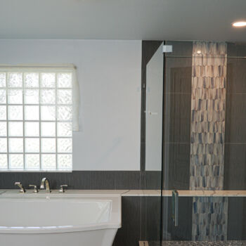 White quartz shelf with grey ribboning for a large bath tub. Large square dark grey wall tile, and grey wood grain floor tiles. Custom shower tiles include dark grey rectangular tiles, vertical ribbon of custom tile also used in niche. Quartz shelf goes all the way across for shower bench. Installation by GWH Flooring in Wimberley, Texas