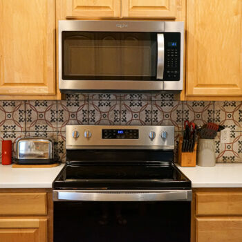 White silestone countertop with red, black, and white decorative pattern backsplash. Grey slate floor tiles. White oak cabinets and black and stainless stove and microwave. Tile Installation in Woodcreek by GWH Flooring store.