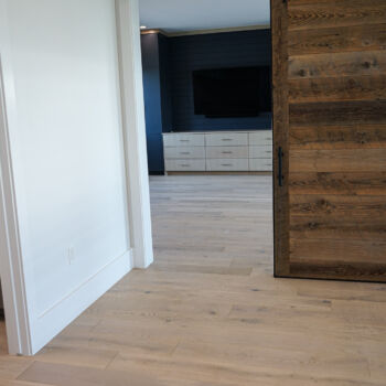 White oak floor installed in a media room with blue cabinets and slatted backing for TV. Room seperated by reclaimed wood barn door. Wood floor continues into bathroom with marble countertop and grey cabinets. Installation of floor and counter top in Driftwood by GWH Flooring company.